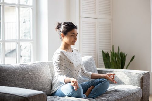 pretty young woman sitting cross-legged on couch at home, meditating - types of meditation