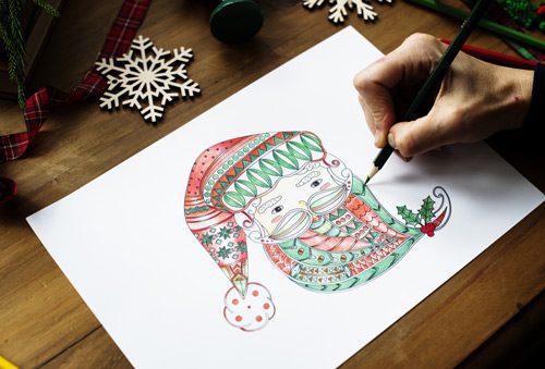 closeup of an adult hand using colored pencils to draw a Santa face on white paper - holiday season